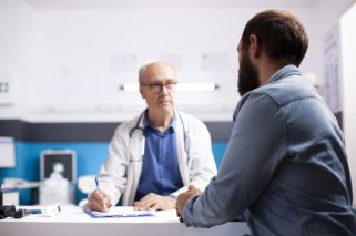 Elderly healthcare specialist in medical office talks with male patient, taking notes on his clipboard. Professional old physician listening to health concerns of young man during clinic appointment.