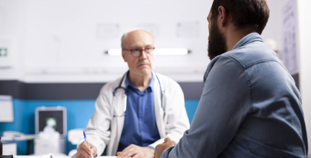 Elderly healthcare specialist in medical office talks with male patient, taking notes on his clipboard. Professional old physician listening to health concerns of young man during clinic appointment.