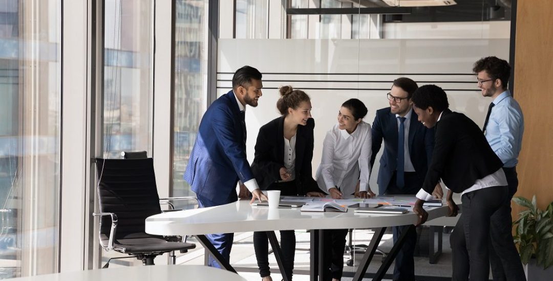 Diverse group of happy employees analyzing reports together, standing at table with papers, working together, discussing marketing, statistic data, talking, brainstorming. Business meeting concept