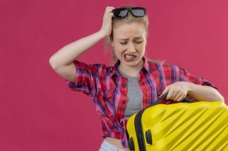 sad traveler young girl wearing red shirt in glasses holding suitcase put her hand on head on isolated pink background