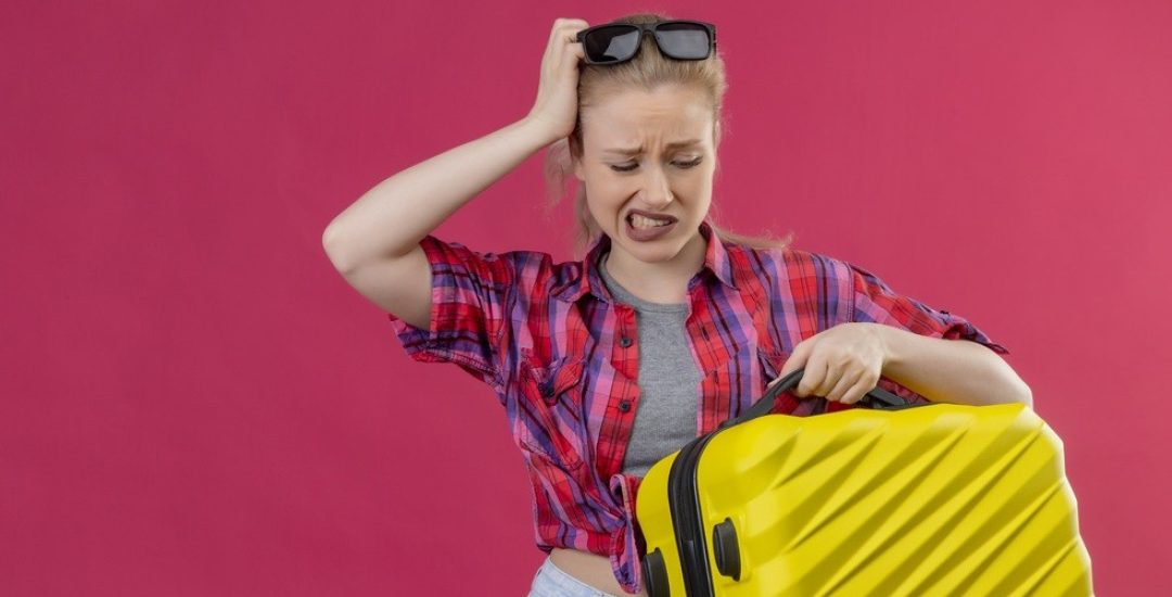 sad traveler young girl wearing red shirt in glasses holding suitcase put her hand on head on isolated pink background