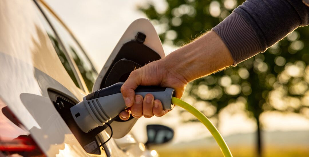 Hand of man inserting a power cord into an electric car for charging ecofriendly vehicle on green landscape