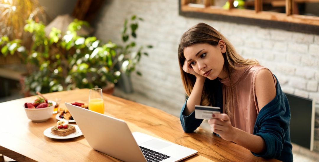 Young woman feeling displeased about debt on her credit card while checking bank account over laptop at home.