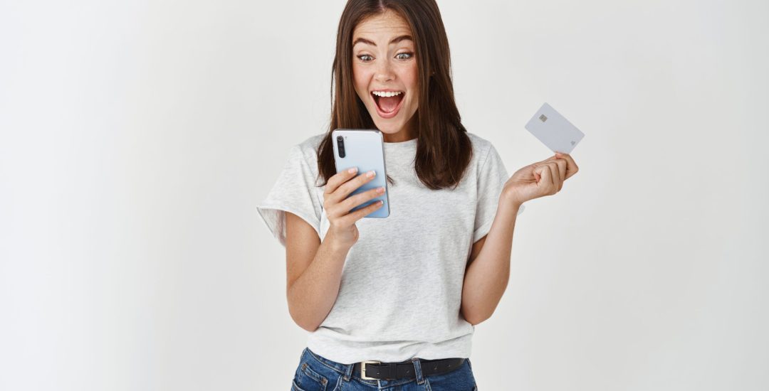 Online shopping and ecommerce. Happy young woman using mobile phone and holding plastic credit card, making purchase, standing over white background.