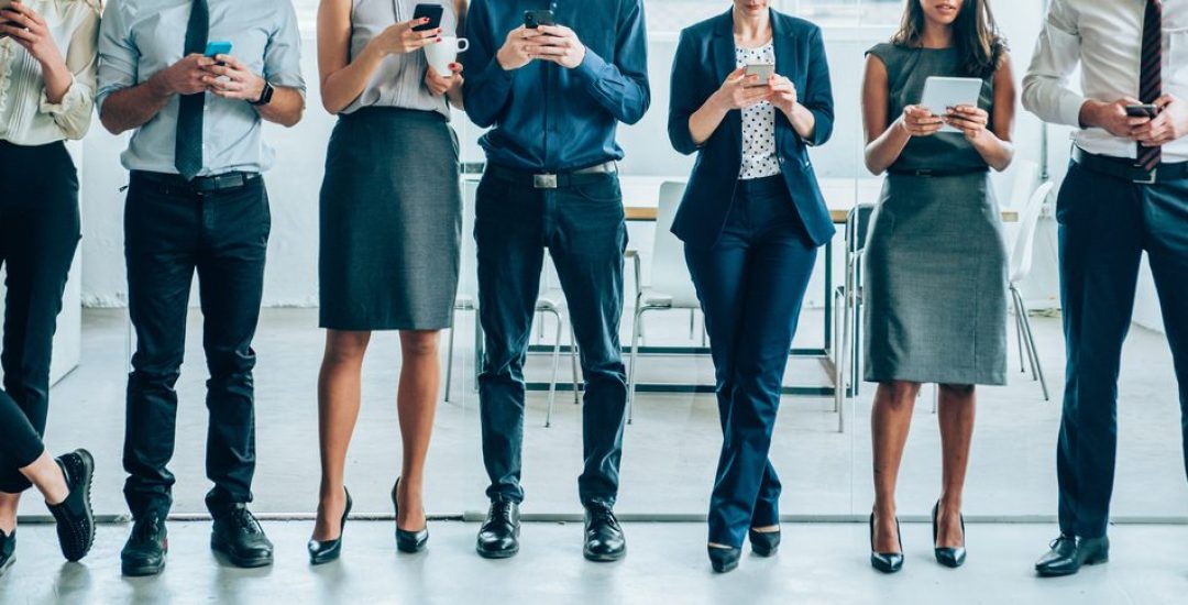 Cropped shot of multi-ethnic group of young business people using mobile devices together in modern office. Corporate business team with tablet and smart phones standing together in a line and having fun in their free time in the office.