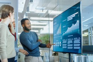 Diverse Team of Young Professionals in Conference Room Have Discussion about Statistics and Graphs Shown on a Presentation TV.