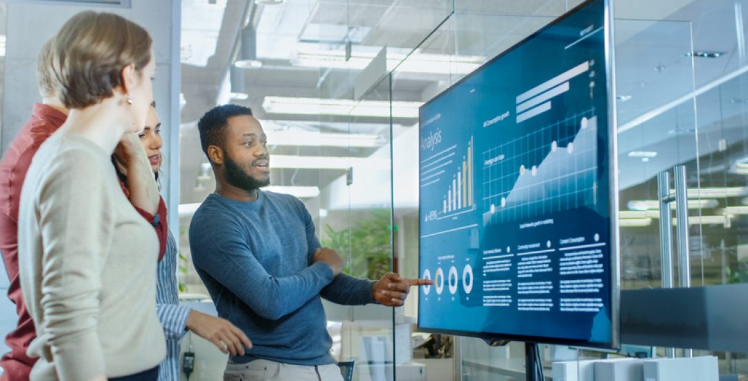 Diverse Team of Young Professionals in Conference Room Have Discussion about Statistics and Graphs Shown on a Presentation TV.
