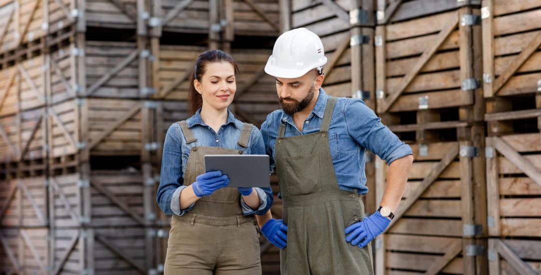 Modern tech for work in warehouse, motion control and logistic. Busy millennial lady in uniform and gloves shows tablet to guy worker in helmet in storage with many wooden containers, free space