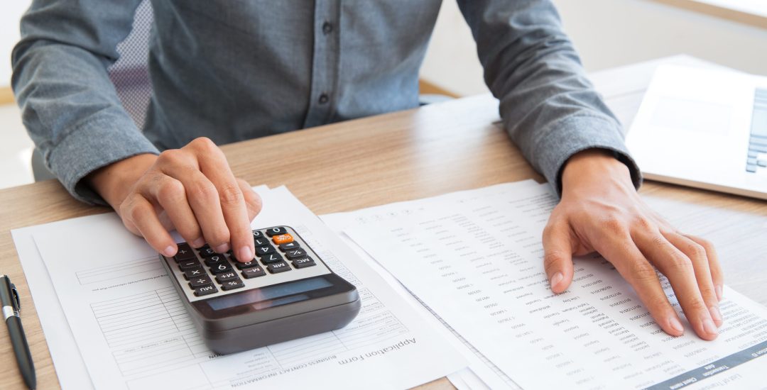Close-up of unrecognizable accountant calculating company income and examining report. He using calculator to check financial data. Man working at table. Investment concept