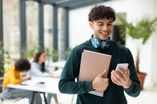 Cheerful,European,Male,Student,Using,Smartphone,And,Holding,Workbooks,Standing