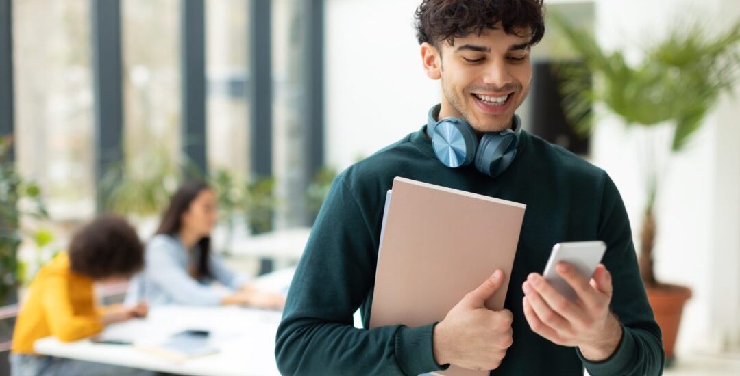 Cheerful,European,Male,Student,Using,Smartphone,And,Holding,Workbooks,Standing