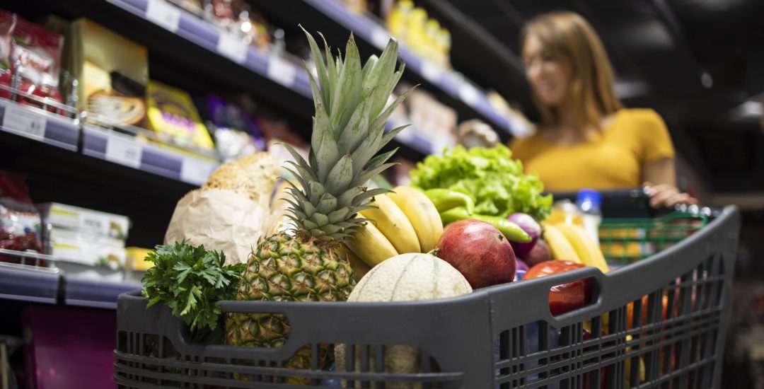 Close up view of shopping cart overloaded with food while in background female person choosing products.