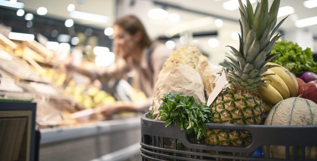 Closeup of shopping cart at supermarket full of food, fruit and vegetables while in background woman taking product off the shelves.