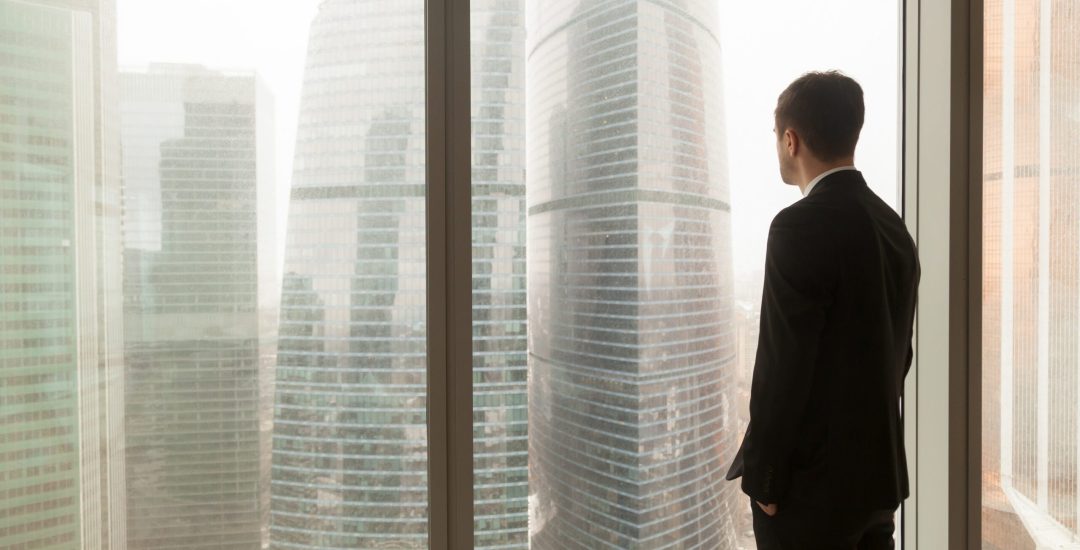 Man in business suit standing with hands in pants pockets looking through window on city landscape with skyscrapers. Businessman thinking about perspectives, dreaming of company success. Back view