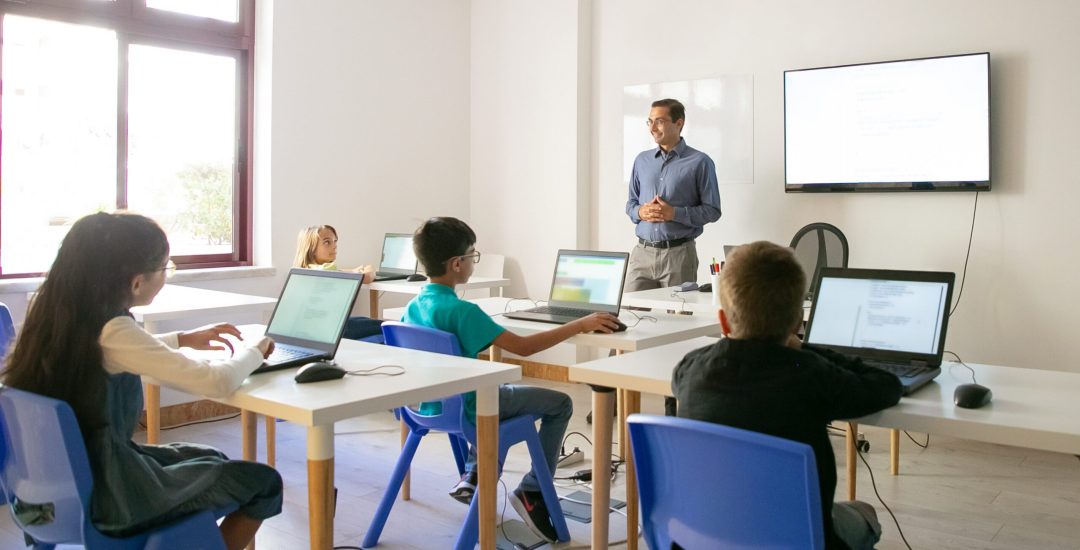 Confident teacher explaining lesson to pupils. Multiethnic children sitting at table in classroom, listening middle-aged man and using laptop computers. Childhood and digital education concept
