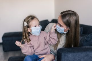 Cute little girl and mother wearing face mask, sitting on bed at home, consoling sad preschool daughter. Concept of coronavirus or COVID-19 pandemic disease symptoms.