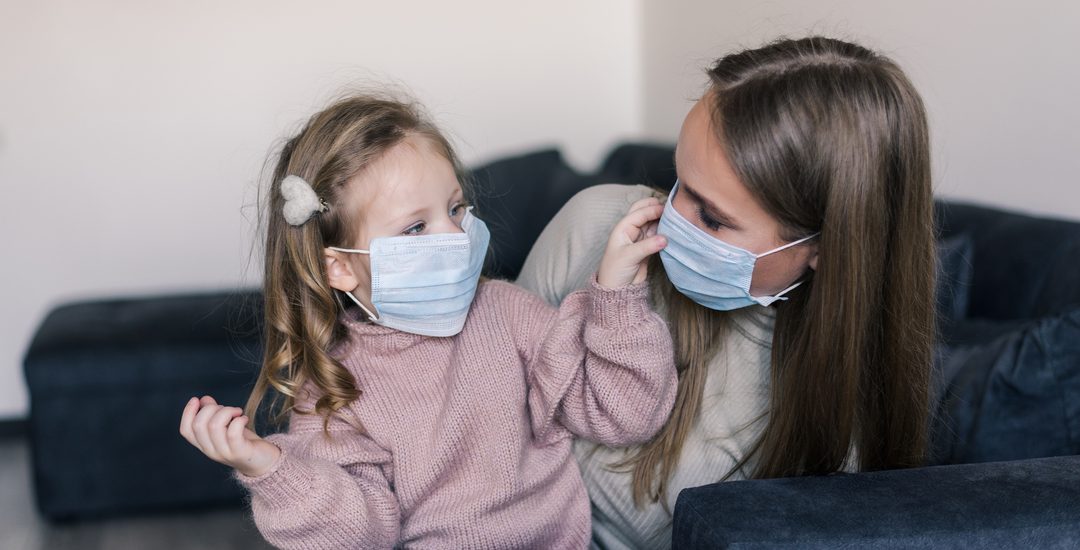 Cute little girl and mother wearing face mask, sitting on bed at home, consoling sad preschool daughter. Concept of coronavirus or COVID-19 pandemic disease symptoms.