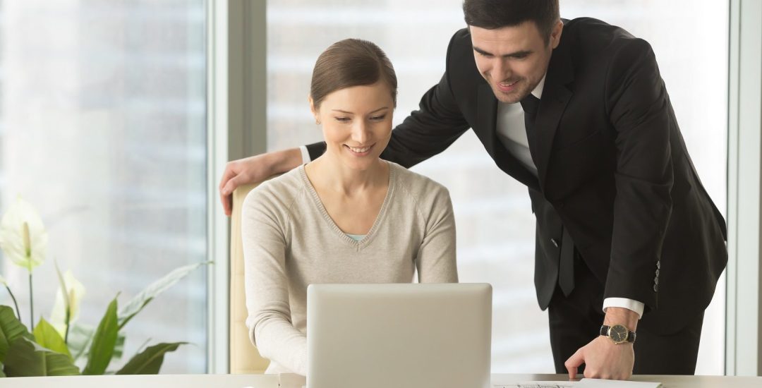 Female and male employees, office colleagues, business partners working together in office. Smiling businesswoman sitting at desk, typing on laptop, businessman standing near by and looking on screen
