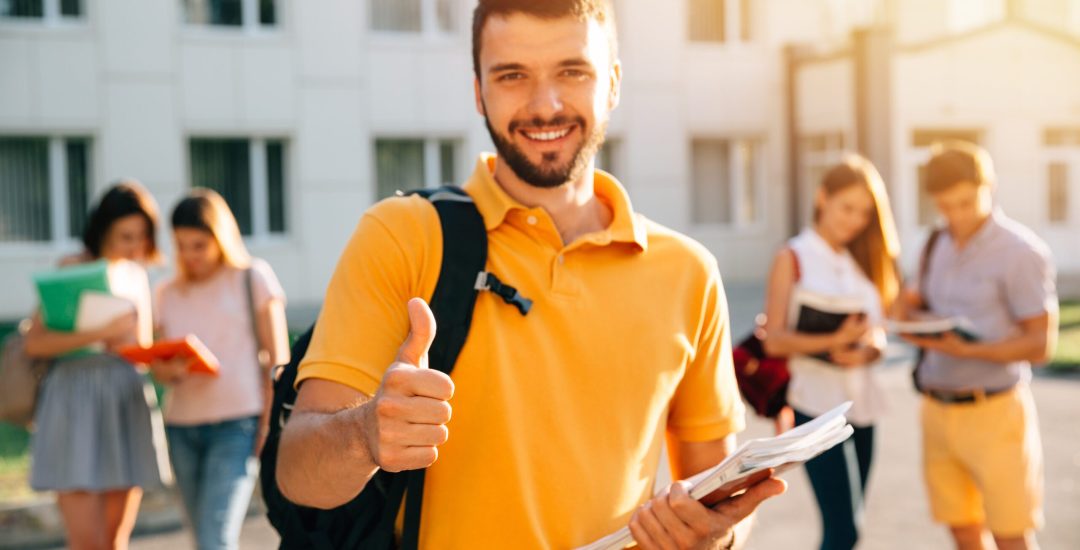 Young attractive smiling student showing thumb up outdoors on campus at the university. Selective focus