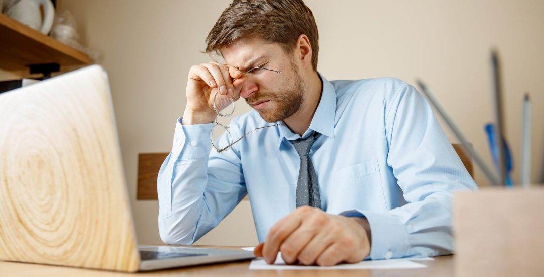 Feeling sick and tired. Frustrated sad unhappy sick young man massaging his head while sitting at his working place in office. The seasonal flu, pandemic influenza, disease prevention concept