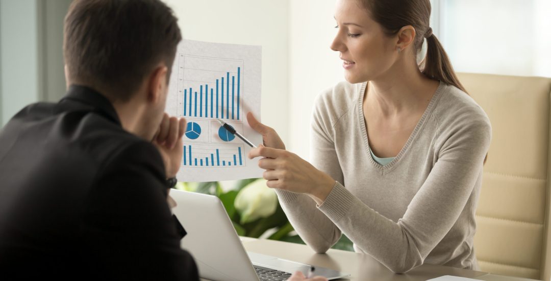 Attractive millennial businesswoman presenting company financial indicators, telling business partner about investment plan, showing document with project positive stats sitting at office desk
