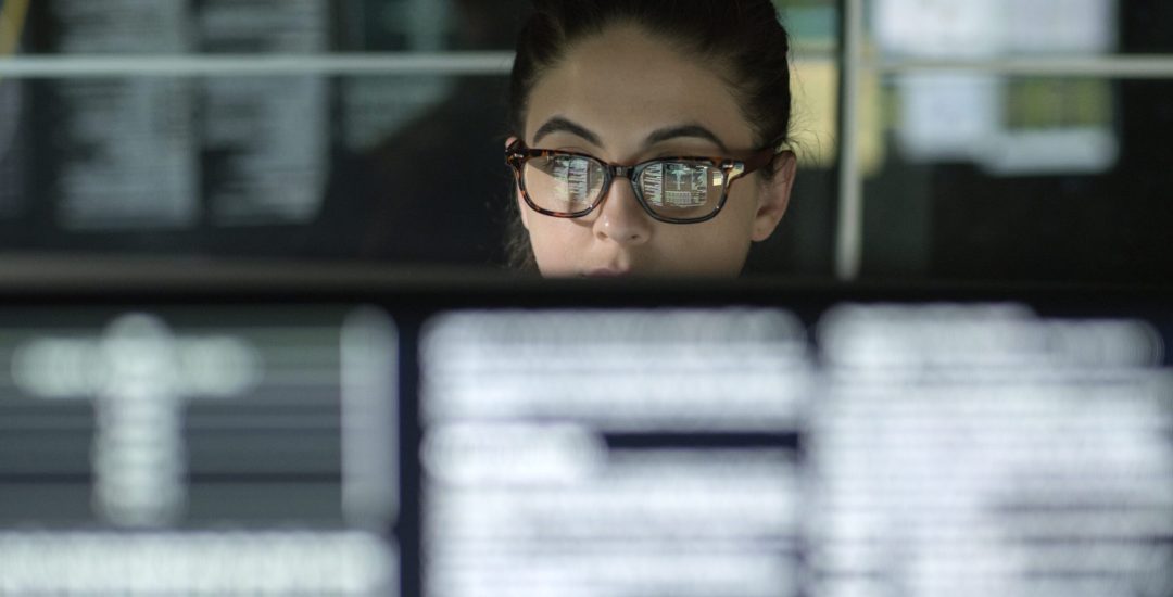 A young woman is surrounded by monitors & their reflections displaying scrolling text & data.