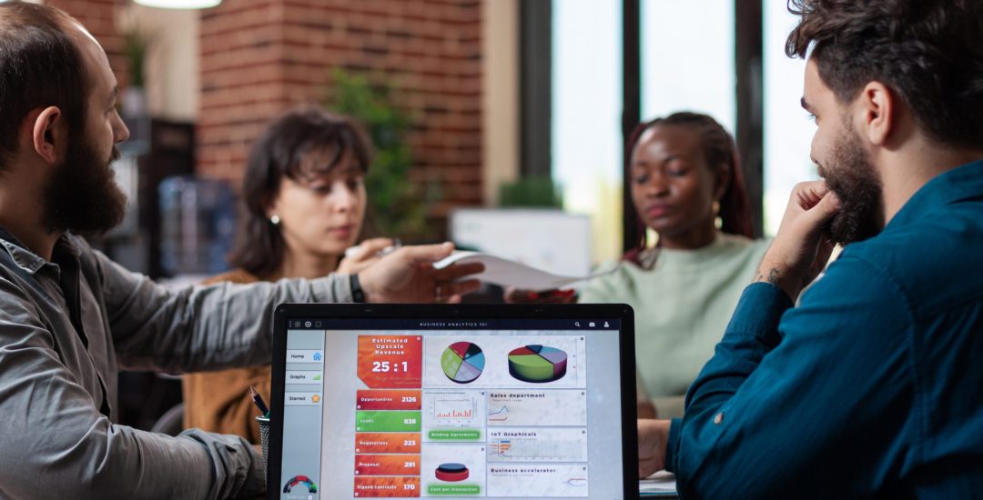 Laptop computer with marketing graphs on screen standing on table in startup office while diverse team sharing papers with company turnover on it. Multiethnic businesspeople analyzing strategy
