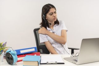 young girl in white shirt and headphones with a microphone looking unwell touching her shoulder feeling pain sitting at the table with folders and laptop over white background