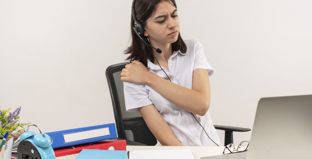 young girl in white shirt and headphones with a microphone looking unwell touching her shoulder feeling pain sitting at the table with folders and laptop over white background