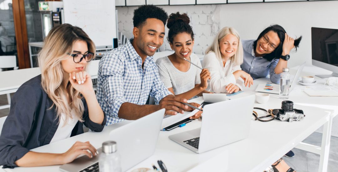 Smiling african student pointing with pencil at laptop screen. Concentrated blonde girl in glasses propping chin with hand while working with computer in office.