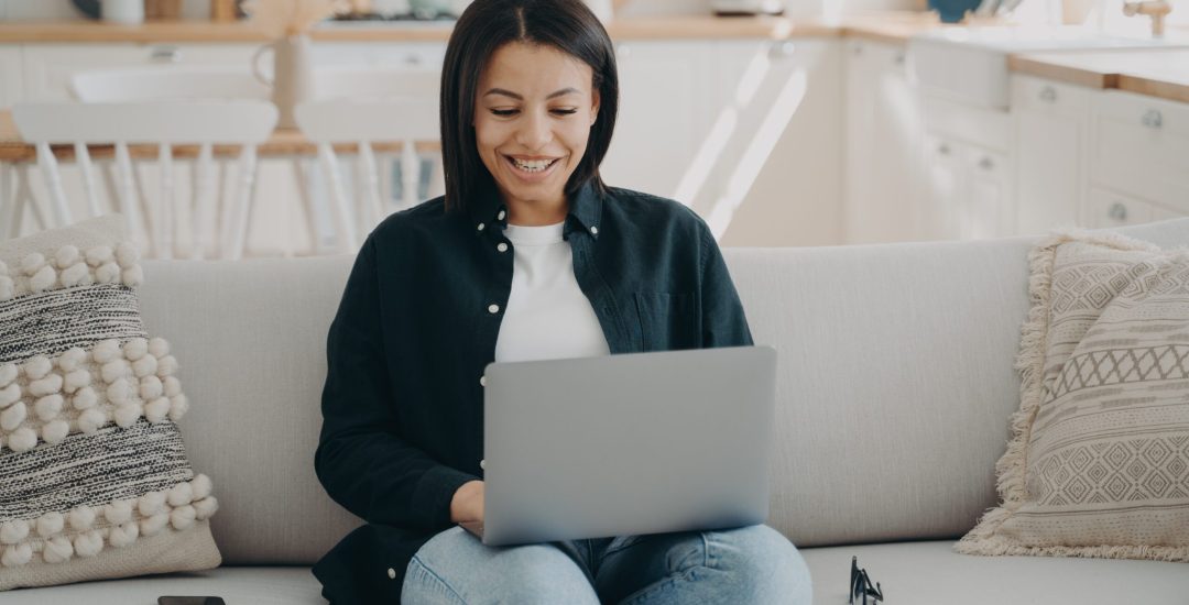 Smiling female shopping on the internet at laptop sitting on couch at home. Happy woman freelancer working on computer, chatting in social networks. Ecommerce, online store advertisement.