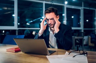 Weary businessman finishing up on work at night. Stressed and tired office worker rubbing his forehead with both of his hands while working behind computer in late hours in office.