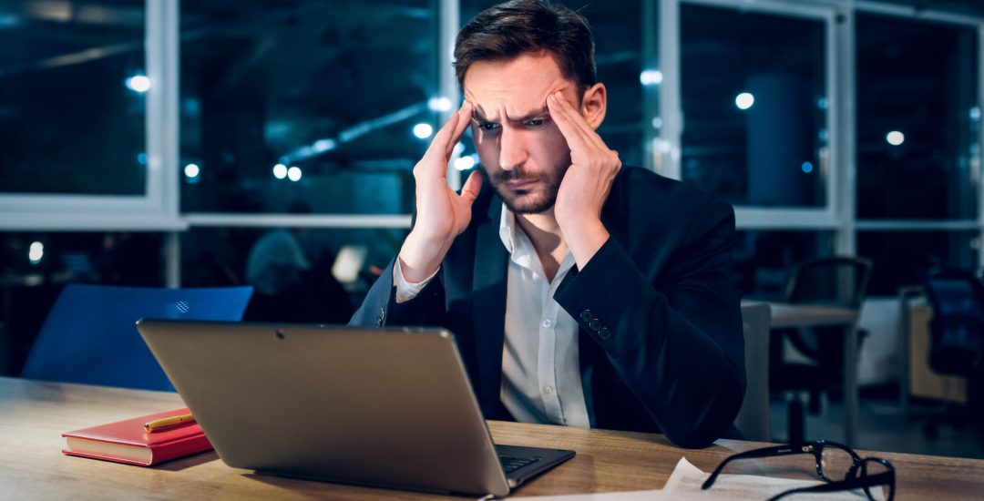 Weary businessman finishing up on work at night. Stressed and tired office worker rubbing his forehead with both of his hands while working behind computer in late hours in office.