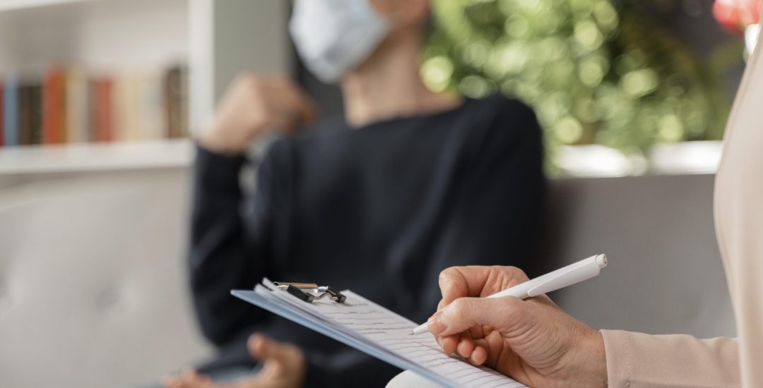 woman-therapist-taking-notes-of-man-in-therapy-office-with-mask