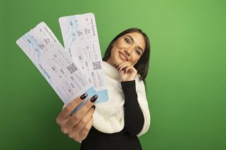 young woman with white scarf showing air tickets looking at camera with smile on face happy and positive standing over green background