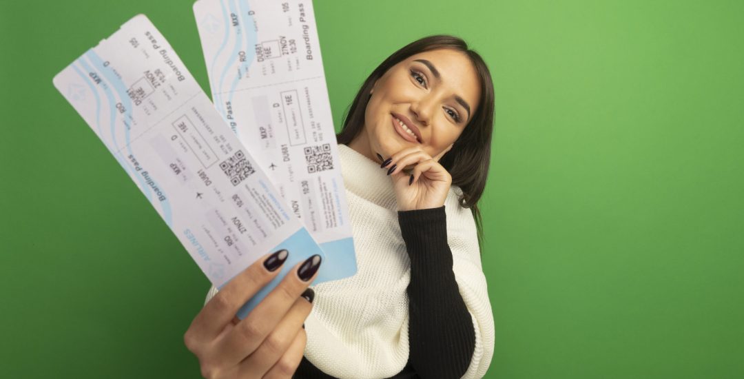 young woman with white scarf showing air tickets looking at camera with smile on face happy and positive standing over green background